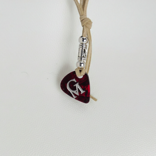 A dark red guitar pick pendant with a silver 'G' monogram, hanging from a tan cord with a silver spacer bead on a textured white background.