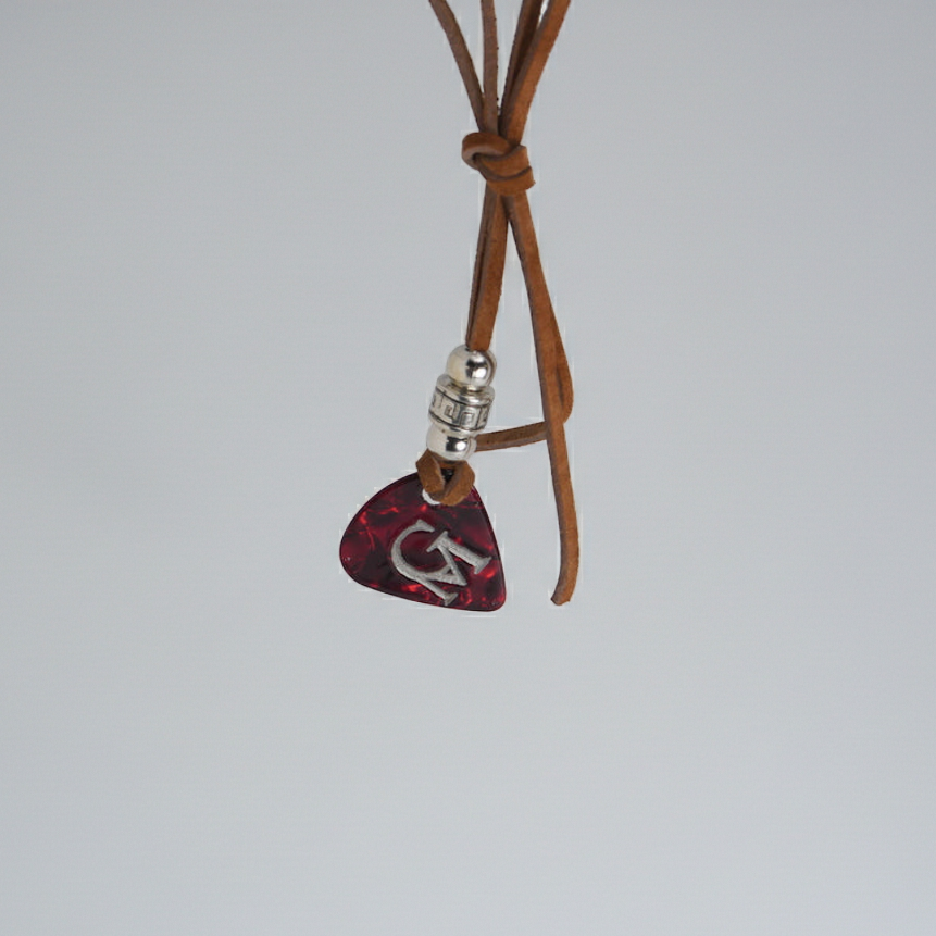 GADHI Founder's Edition bracelet: brown leather cord with a red and black guitar pick engraved with the silver GM emblem, on a light gray background.