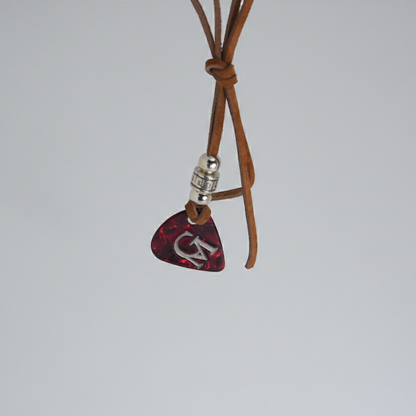 GADHI Founder's Edition bracelet: brown leather cord with a red and black guitar pick engraved with the silver GM emblem, on a light gray background.
