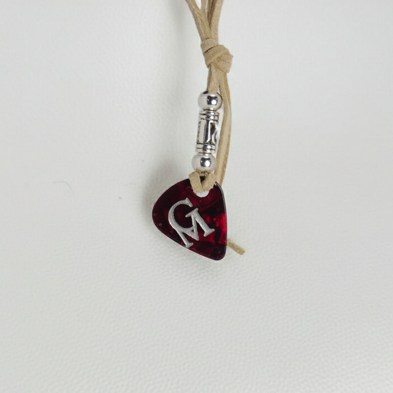 A dark red guitar pick pendant with a silver 'G' monogram, hanging from a tan cord with a silver spacer bead on a textured white background.